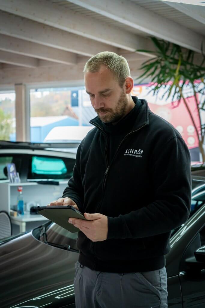 Ein Mann im schwarzen Sakko erstellt am Tablet neben einem Auto ein Unfallgutachten in einem modernen Showroom oder Büro