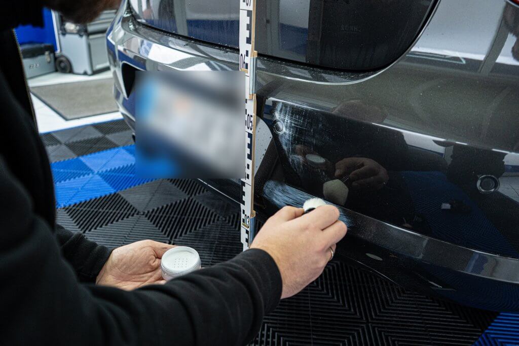 Eine Person prüft und misst Kratzer an einer Autostoßstange mit einem Lineal für ein Unfallgutachten, das Nummernschild ist unkenntlich