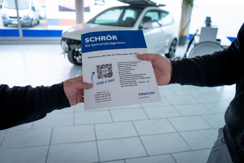 Zwei Personen tauschen im Autohaus eine Broschüre des Schrör Kfz-Sachverständigenbüros, im Hintergrund steht ein weißes Auto