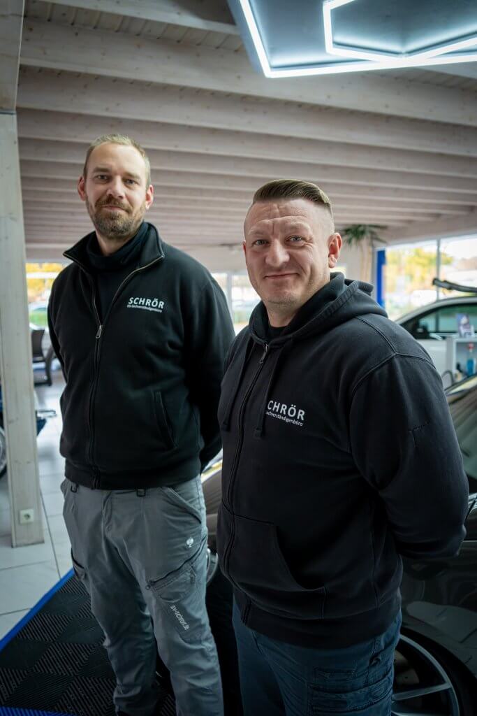 Zwei Männer in schwarzen Kapuzenpullis stehen lachend nebeneinander in einem hellen, modernen Autohaus zwischen mehreren Fahrzeugen